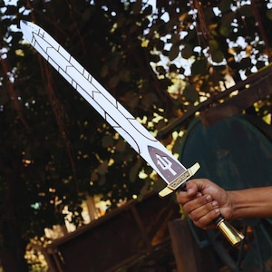 May include: A white and black sword with a brown hilt and gold accents. The sword features a trident symbol and text on the hilt. The sword is held in a hand against a blurred background of green foliage.