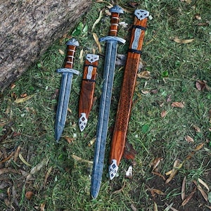 May include: Three swords with wooden sheaths. The swords have silver accents and are laid out on a bed of green grass.