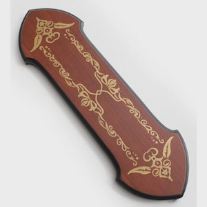 May include: A rectangular wooden plaque with pointed ends and a dark brown finish. The surface features a gold-colored decorative filigree pattern with symmetrical floral motifs at the top and bottom, framed by a thin black border along the edges.