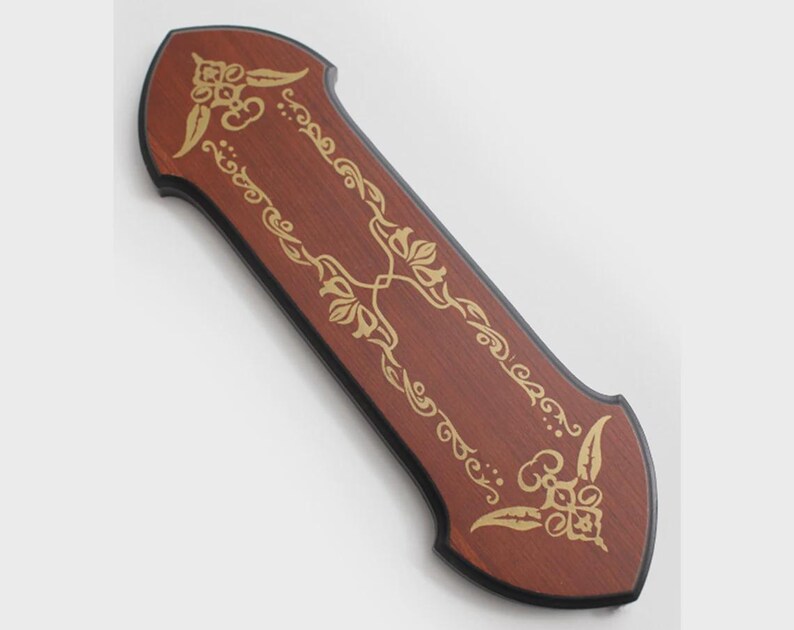 May include: A rectangular wooden plaque with pointed ends and a dark brown finish. The surface features a gold-colored decorative filigree pattern with symmetrical floral motifs at the top and bottom, framed by a thin black border along the edges.