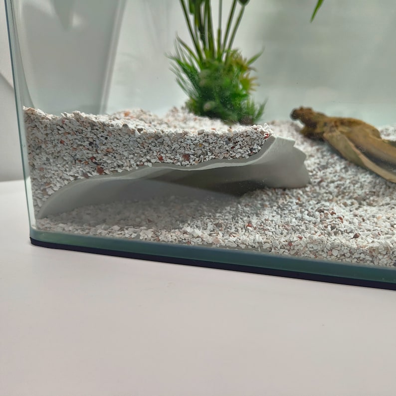 Pu&ograve; includere: Un acquario in vetro trasparente con base nera, riempito di ghiaia colorata chiara e una struttura bianca curva. Una piccola pianta verde e un pezzo di legno galleggiante sono visibili all'interno, suggerendo un habitat acquatico.