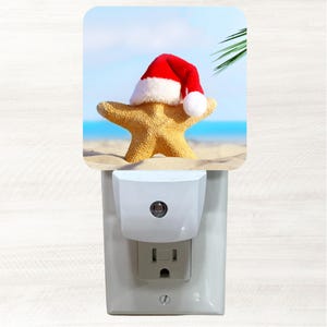 May include: A white plastic night light featuring a square decorative plate with a printed image of a yellow starfish wearing a red and white Santa hat on a sandy beach with a blue sky and ocean background and a green palm leaf in the corner.