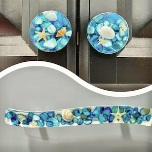May include: Decorative cabinet knobs and a handle featuring a beach theme. The knobs are round, with blue and turquoise stones, seashells, and starfish encased in clear resin. The handle has a similar design, with a rectangular shape.
