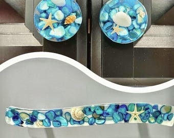 May include: Decorative cabinet knobs and a handle featuring a beach theme. The knobs are round, with blue and turquoise stones, seashells, and starfish encased in clear resin. The handle has a similar design, with a rectangular shape.