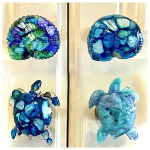 May include: Four decorative cabinet knobs in the shape of a seashell, a turtle, and two different styles of a turtle. The knobs are made of clear resin with blue and green glass pieces embedded in the resin.