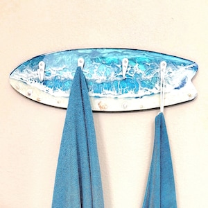 May include: A surfboard-shaped wall hook with a blue and white ocean wave design. The hooks are white and hold two blue towels. The board is decorated with seashells and has a glossy finish, creating a coastal theme.