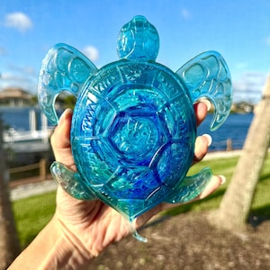 May include: A translucent blue turtle figurine with intricate details. The sea turtle has a textured shell and flipper-like wings. The figurine is held up against a backdrop of a blue sky and water.