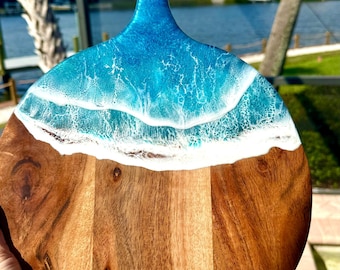 May include: A round wooden cutting board with a blue and white epoxy resin design resembling ocean waves. The board has a handle and is perfect for serving cheese, charcuterie, or other appetizers.