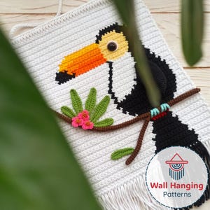 May include: White crochet wall hanging with a toucan design. The toucan has a yellow head, orange and black beak, and black body. A branch with green leaves and pink flowers is below the toucan. The wall hanging has a fringe at the bottom.