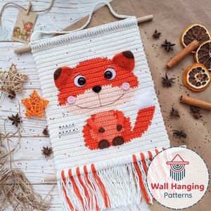 Könnte beinhalten: Handgefertigter Wandbehang aus weißem Häkelstoff mit einem lächelnden orangefarbenen Fuchs. Der Fuchs hat braune Akzente und rosafarbene Wangen. Der Wandbehang trägt den Text "MAGIC MOMENT" und hat einen orange-weißen Fransenabschluss. Wanddekoration.