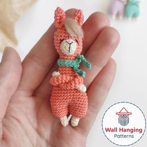 May include: A handmade, coral-colored crochet llama with a light brown fringe, a turquoise scarf, and white accents. The llama is wearing pants and has a smiling face. The image includes a logo for Wall Hanging Patterns.