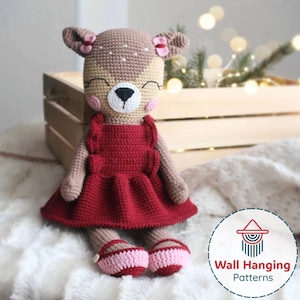 May include: A handmade crochet deer doll with a tan head and body, wearing a red dress and pink shoes. The deer has a white muzzle, black nose, and pink cheeks. The ears are tan with pink bows. The doll is sitting on a white surface.