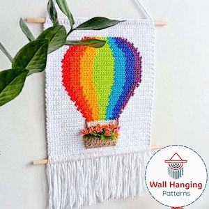 May include: A handmade wall hanging featuring a colorful rainbow hot air balloon design. The balloon is crafted with vibrant red, orange, yellow, green, blue, and purple yarn. A woven basket with flowers is attached below. The wall hanging is white with fringe.