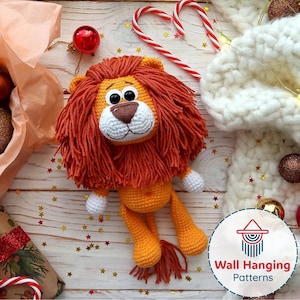 Op de afbeelding: Een handgemaakte gehaakte leeuwenpop met een oranje manen, lichaam en poten, en een wit gezicht en poten. De leeuw ligt op een houten oppervlak versierd met kerstversieringen, zuurstokken en stervormige confetti.