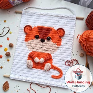 May include: A crochet wall hanging depicting an orange tiger on a white background. The tiger has black eyes and a light beige muzzle. The rectangular wall hanging has wooden dowels at the top and bottom, and a white cord for hanging. The design is handmade.
