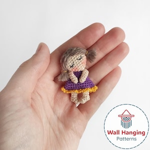 May include: A handmade crochet doll with a purple dress and yellow trim, held in a hand. The doll has closed eyes, rosy cheeks, and brown hair in pigtails. The doll is a miniature craft item.