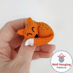 Pode incluir: Estatueta de raposa laranja de crochê, com detalhes dourados, nariz preto e olhos fechados. A raposa tem uma cauda com ponta branca e está enroscada a dormir. Objeto decorativo feito à mão.