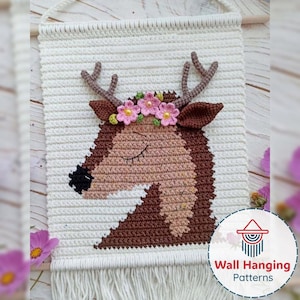 Pode incluir: Tapeçaria de parede de crochê com a cabeça de um veado com olhos fechados, chifres marrons e uma coroa de flores. O veado é feito em tons de marrom e bege, sobre um fundo branco com franjas. Pendurado em uma vara de madeira.
