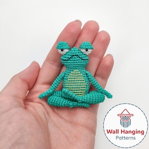 May include: A small, turquoise crochet frog in a seated yoga pose. The frog has large, round eyes and a yellow stitched pattern on its belly. The frog is held in a hand, with a white background.