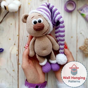Pode incluir: Boneca de urso de crochê feita à mão, usando um gorro listrado roxo e branco e botas roxas. O urso tem um nariz marrom, olhos pretos e uma expressão sorridente. A boneca é segurada nas mãos de uma pessoa. Um brinquedo macio e fofo.