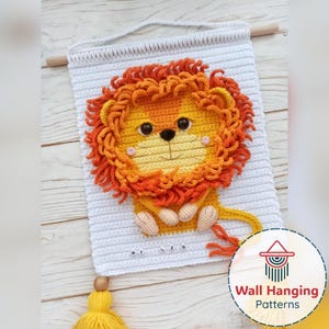 May include: A handmade crochet wall hanging featuring a lion design. The lion has a yellow and orange mane, a yellow face, and a white background. The wall hanging is finished with a wooden dowel and a yellow tassel.