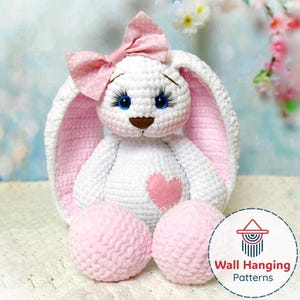 Puede incluir: Un conejo blanco de ganchillo hecho a mano con detalles en rosa. El conejo tiene ojos azules, un lazo rosa y un corazón rosa en el pecho. Dos bolas rosas en la base. Un juguete suave.