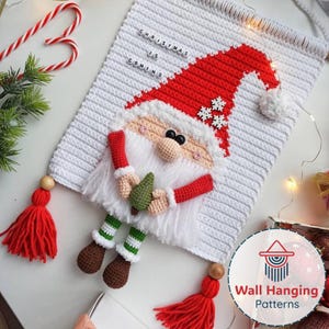 May include: A crocheted Christmas wall hanging with a Santa Claus design. The Santa has a red hat, white beard, and holds a small green tree. The wall hanging is white with the text "CHRISTMAS IS COMING". Red tassels and a candy cane are also visible.