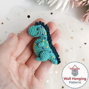 May include: A turquoise crochet dinosaur with a dark blue spine and yellow and purple accents. The handmade dinosaur has a smiling face and is likely a toy or decorative item. The dinosaur is held in a hand.