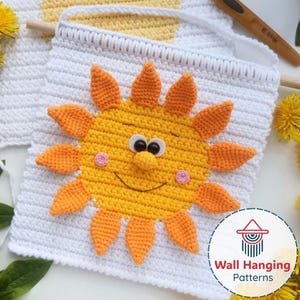 Op de afbeelding: Handgemaakte gehaakte wanddecoratie met een vrolijk zonontwerp. De zon is geel met oranje stralen, wiebelogen en roze wangen, tegen een witte achtergrond. De wanddecoratie is bevestigd aan een houten stok.