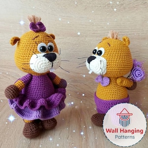 May include: Two handmade crochet beaver dolls. One beaver wears a purple dress and the other holds a purple flower bouquet. Both have yellow fur, white teeth, and black eyes. The dolls are on a light brown surface.