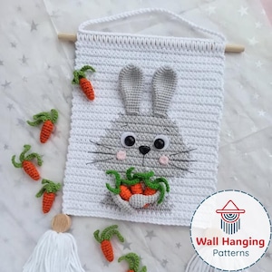May include: A white crochet wall hanging with a grey bunny face, black eyes, pink cheeks, and a bowl of carrots. Several crochet carrots with green tops are scattered around the hanging. The wall hanging is suspended by a wooden dowel and white cord.