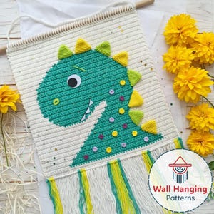 May include: A cream-coloured, crocheted wall hanging with a green dinosaur design. The dinosaur has yellow and green accents, a large eye, and decorative spots. The hanging has a fringe at the bottom and a wooden dowel at the top. The text "Wall Hanging Patterns" is visible.