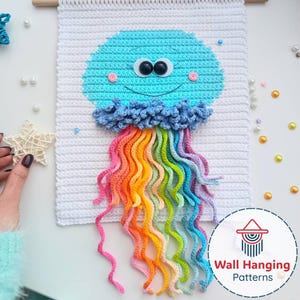 Pode incluir: Um pendurador de parede de crochê feito à mão com um design de água-viva sorridente. A água-viva tem uma cabeça azul clara, uma gola azul e tentáculos de arco-íris. O fundo é branco e a peça é montada em um bastão de madeira. O texto "Wall Hanging Patterns" está no canto inferior direito.