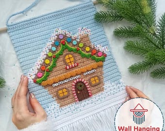 Op de afbeelding: Een gehaakte wandhanger met een peperkoekhuisontwerp. Het huis is bruin met kleurrijke snoepdecoraties, een groene slinger en een besneeuwd dak. De achtergrond is lichtblauw en de onderkant heeft witte franjes.