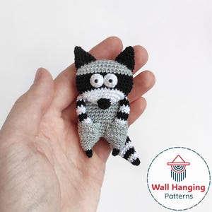 May include: A crocheted raccoon figurine in gray, black, and white. The handmade animal has a striped tail and a bandit mask design. The figurine is held in a hand. The image also includes the text "Wall Hanging Patterns".