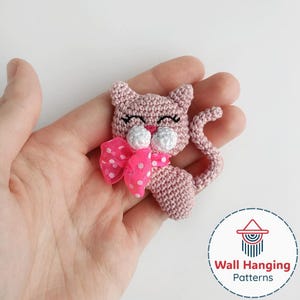 May include: A crocheted pink cat with a pink polka dot bow. The cat has black eyelashes and white accents. The cat's tail is curled. The image includes the text "Wall Hanging Patterns" in a circular logo.