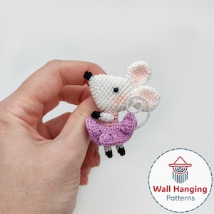 Puede incluir: Una figura de ratón de ganchillo hecha a mano con cuerpo blanco, orejas rosas y falda morada. El ratón tiene ojos y pies negros. La figura se sostiene en una mano. La imagen incluye el texto "Wall Hanging Patterns".