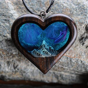 May include: Heart-shaped pendant with a dark wood frame. The center features a blue and purple design with a mountain range at the bottom. The pendant hangs from a black cord with a silver-colored clasp.