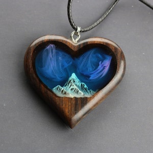 May include: Heart-shaped wooden pendant with a mountain scene. The pendant features a dark wood frame with a resin interior depicting a mountain range and swirling blue and purple clouds. The pendant hangs from a black cord.
