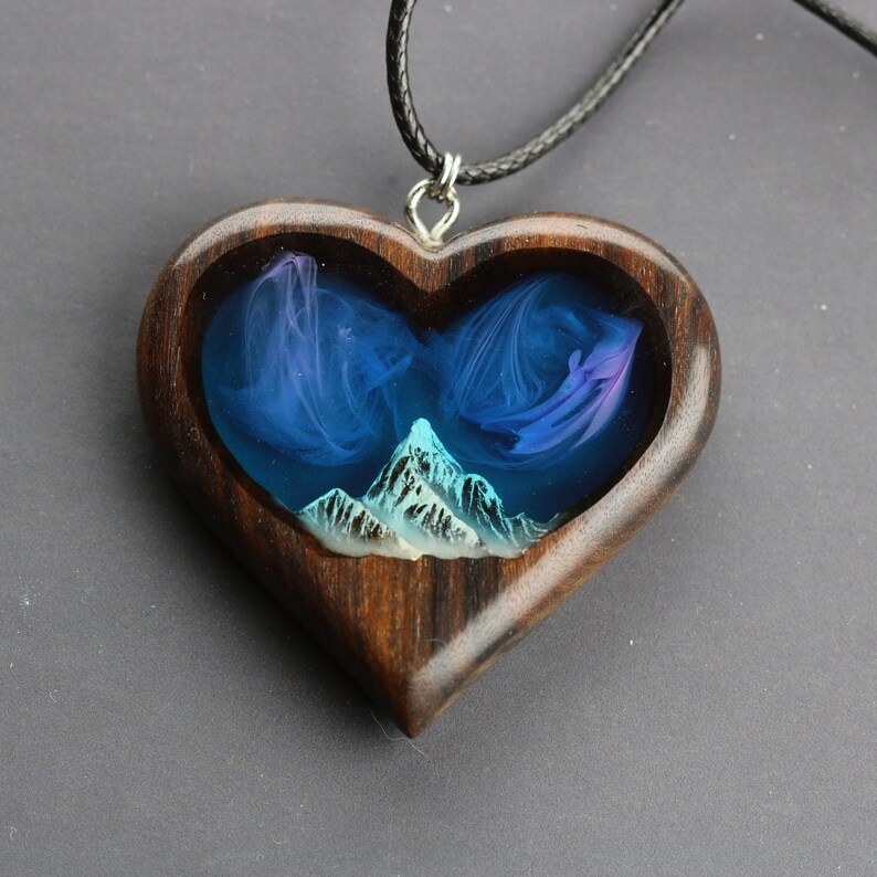May include: Heart-shaped wooden pendant with a mountain scene. The pendant features a dark wood frame with a resin interior depicting a mountain range and swirling blue and purple clouds. The pendant hangs from a black cord.