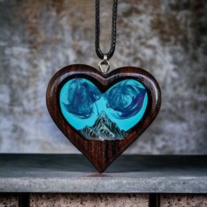May include: Heart-shaped wooden pendant with a turquoise resin center featuring a mountain scene. The pendant has a dark brown wood frame and hangs from a black cord. The resin art has blue and purple swirls.