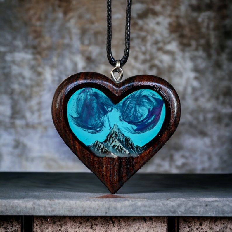 May include: Heart-shaped wooden pendant with a turquoise resin center featuring a mountain scene. The pendant has a dark brown wood frame and hangs from a black cord. The resin art has blue and purple swirls.