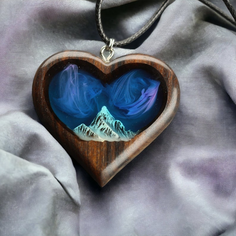 May include: A heart-shaped pendant with a dark wood frame. The center features a blue and purple swirling design with a mountain range at the bottom. The pendant hangs from a black cord.