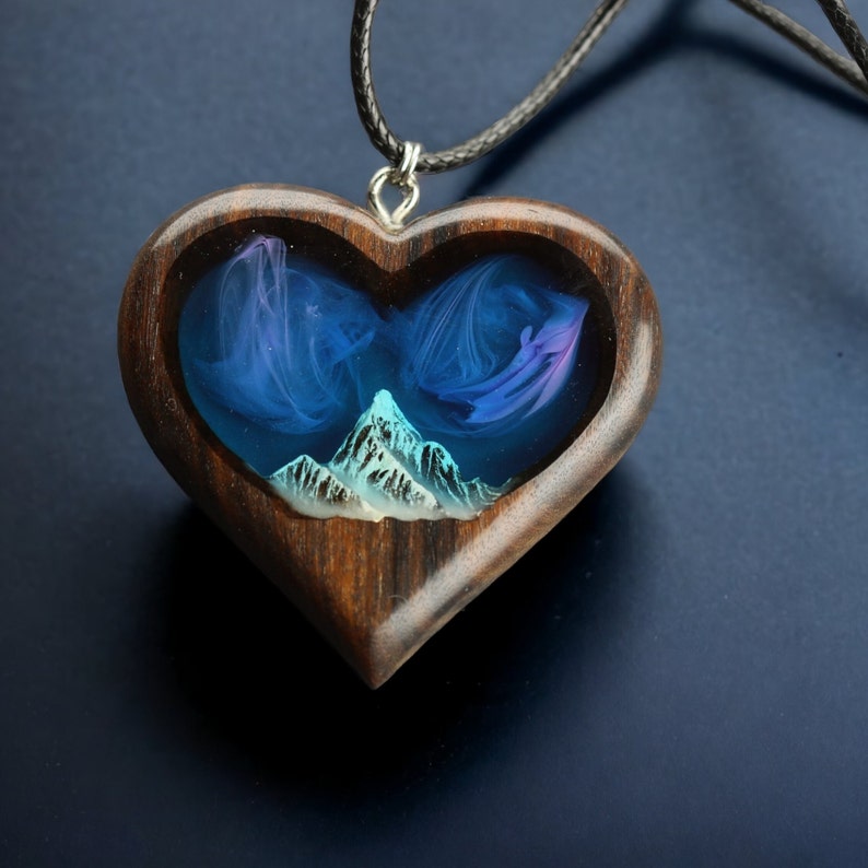 May include: Heart-shaped wooden pendant with a mountain scene. The pendant features a dark wood frame with a blue and purple resin interior depicting a mountain range. The pendant hangs from a black cord.