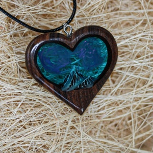 May include: Heart-shaped wooden pendant with a landscape scene. The pendant features a dark wood frame, enclosing a blue and green scene with mountains and swirling clouds. The pendant hangs from a black cord.