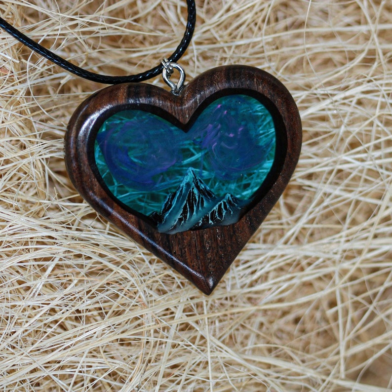 May include: Heart-shaped wooden pendant with a landscape scene. The pendant features a dark wood frame, enclosing a blue and green scene with mountains and swirling clouds. The pendant hangs from a black cord.