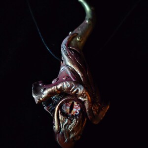 Könnte beinhalten: Eine handgefertigte Anhänger-Halskette mit Fantasy-Thema. Der Anhänger hat ein detailliertes, mehrfarbiges Design mit einem gebogenen Horn, einem Auge und dem Wort "FIRE" in weißer Schrift. Der Anhänger hängt an einer schwarzen Kordel.