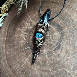 Op de afbeelding: Een zwarte en gouden ketting met een hanger met een blauw oog en ingewikkelde details. De hanger is puntig en hangt aan een zwart koord.