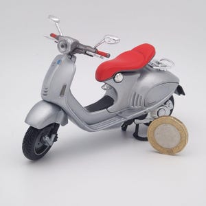 May include: A silver toy scooter with a red seat. The scooter has black tyres, a silver handlebar, and a small windshield. A coin is placed next to the scooter for scale.