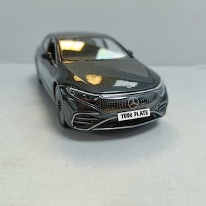 May include: A dark grey model car with a metallic finish. The car features a distinctive front grille with the Mercedes-Benz emblem and the text "YOUR PLATE" on the number plate. The car's design is modern and streamlined.